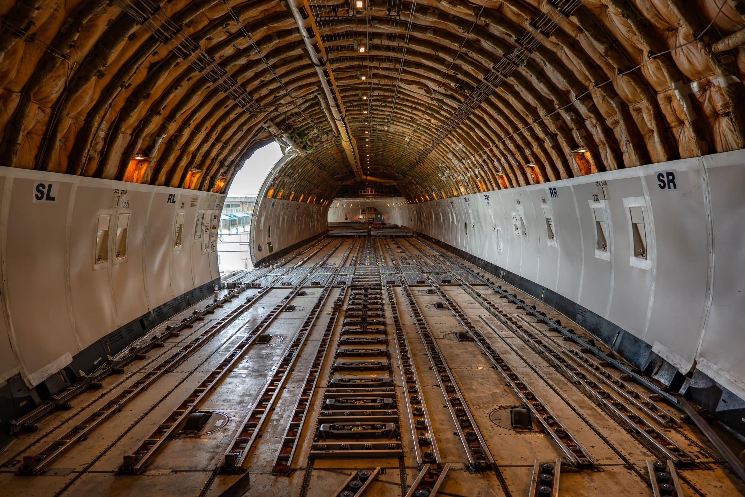 The main deck of a freighter aircraft, looking aft along the roller floor and cargo tracks toward the open main-deck door.